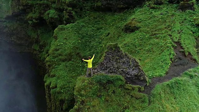 慢镜头男子举起手臂站在悬崖上瀑布前庆祝成功的旅行和冒险