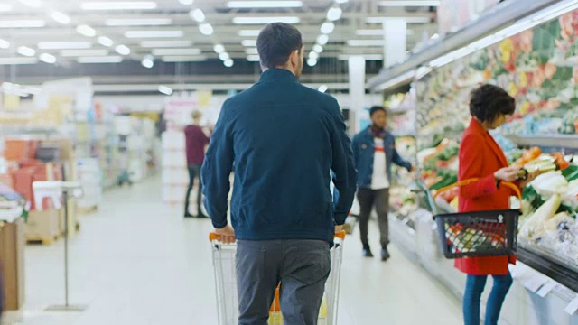 在超级市场:推着购物车的人通过商店的新鲜农产品区。商店与许多顾客购物。后面的视图拍摄。