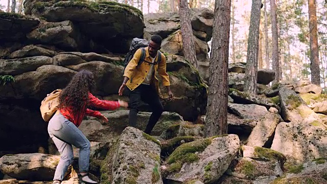 一对年轻的多种族夫妇正在爬山，一名非洲裔美国男子正在帮助一名妇女，向她伸出手并把她拉起来，人们拿着背包。