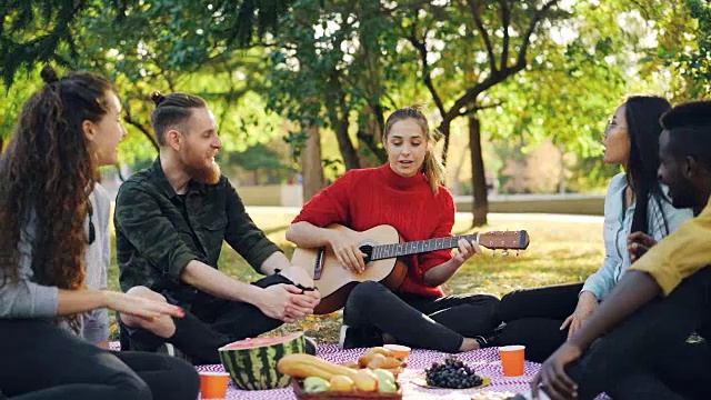 詳情圖-當(dāng)美麗的年輕女子在公園野餐時，歡快的人們唱歌和大笑的慢鏡頭。音樂，休閑和樂趣的概念。