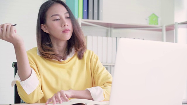 美丽的年轻微笑的亚洲妇女工作笔记本电脑在客厅在家里。亚洲商务女性写笔记本文件财务和计算器在家庭办公室。享受居家时光的概念。