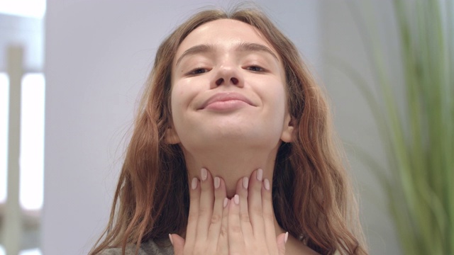 详情图-快乐的女人一边用手做面部按摩，一边对着浴室的镜子