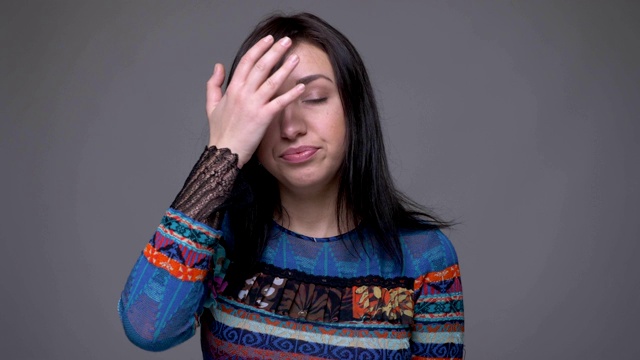 近距离拍摄的成年白人黑发女性戴着她的头尴尬被激怒看与灰色的背景孤立的相机