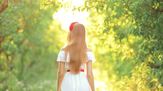 女孩穿着漂亮的白色连衣裙在公园里散步。晴朗的夏日。后视图。