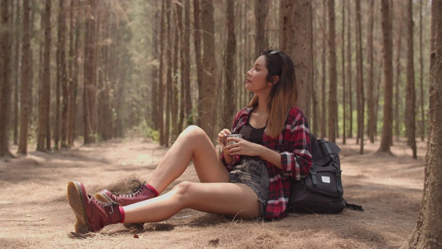 在森林中徒步旅行的亚洲女徒步旅行者。年轻快乐的背包女孩坐着休息喝着水杯后走着旅行，去大自然探险旅行，在暑假爬山度假的概念。缓慢的运动。