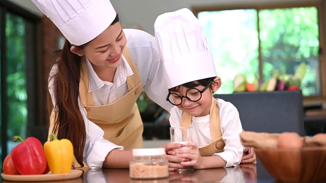 美丽的亚洲女人和可爱的小男孩戴着眼镜一起准备在家里的厨房做饭。生活方式和家庭。儿子不喜欢牛奶，拿着牛奶杯做鬼脸。食物和饮料