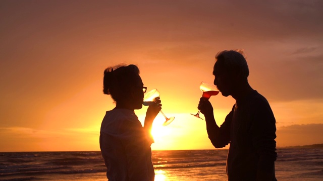 剪影亚洲夫妇老年人退休放松在海滩和喝葡萄酒或香槟，拥抱在日落蜜月家庭一起幸福的人的生活方式，慢镜头