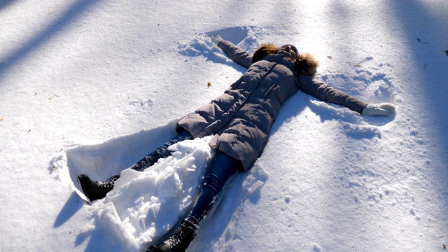 年轻的女孩躺在白雪覆盖的草地上做天使。快乐的女人在雪地里玩，享受冬天的时光。快乐的女士在户外玩。寒假的概念。慢镜头