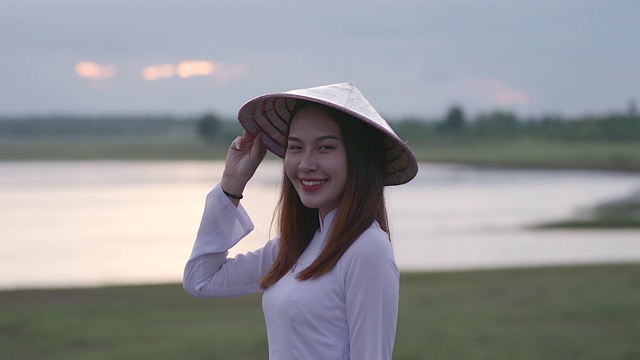 一位美丽的年轻女子穿着一件白色的连衣裙，拿着一顶竹编织的帽子。站着对着镜头笑，心情很好
