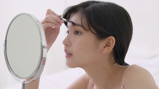 美丽的年轻亚洲女人用化妆眉刷，美丽的亚洲女孩用化妆品为化妆风格的镜子在家里，时尚的女性镊子与自然，生活方式的概念。