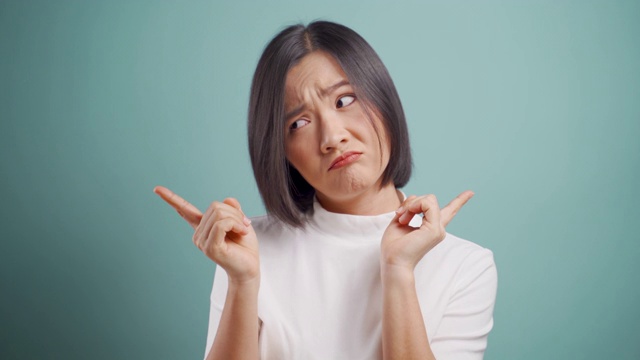 亚洲女性感到困惑的手指左或右看着孤立的摄像头在蓝色背景。4 k的视频。情感的民主观。