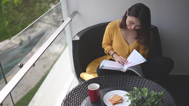 詳情圖-一位亞洲華裔美女中午在她的陽臺(tái)上看書，放松，享受她的數(shù)字排毒時(shí)間，喝著咖啡，吃著餅干當(dāng)點(diǎn)心