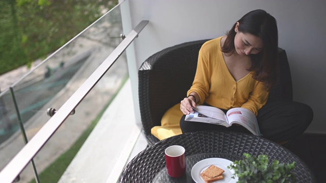 詳情圖-一位亞洲華裔美女中午在她的陽臺(tái)上看書，放松，享受她的數(shù)字排毒時(shí)間，喝著咖啡，吃著餅干當(dāng)點(diǎn)心