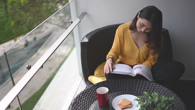 詳情圖-一位亞洲華裔美女中午在她的陽臺(tái)上看書，放松，享受她的數(shù)字排毒時(shí)間，喝著咖啡，吃著餅干當(dāng)點(diǎn)心
