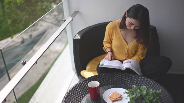詳情圖-一位亞洲華裔美女中午在她的陽臺(tái)上看書，放松，享受她的數(shù)字排毒時(shí)間，喝著咖啡，吃著餅干當(dāng)點(diǎn)心