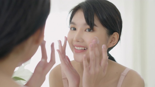 美丽的年轻亚洲女人微笑看着镜子检查脸和皮肤护理霜和化妆品为年轻化和卫生，美丽的女孩快乐清洁面部与乳液和健康。