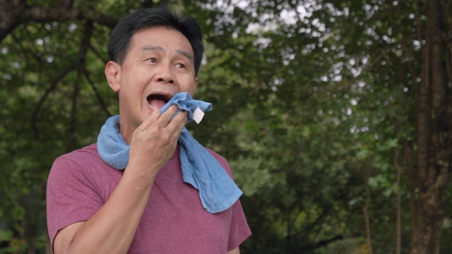 手持拍摄和中拍摄:亚洲老人早上在公园跑步后休息和喝水。他用毛巾擦去锻炼后的汗水。他做完瑜伽和太极运动后很累。保健和运动概念。