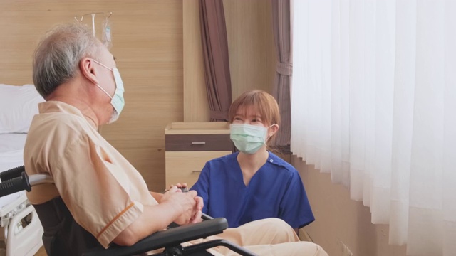 在医院照顾坐在轮椅上的成熟男性病人的亚洲护士。年轻妇女和老人戴上外科口罩，以预防covid - 19大流行。女孩对老人微笑。