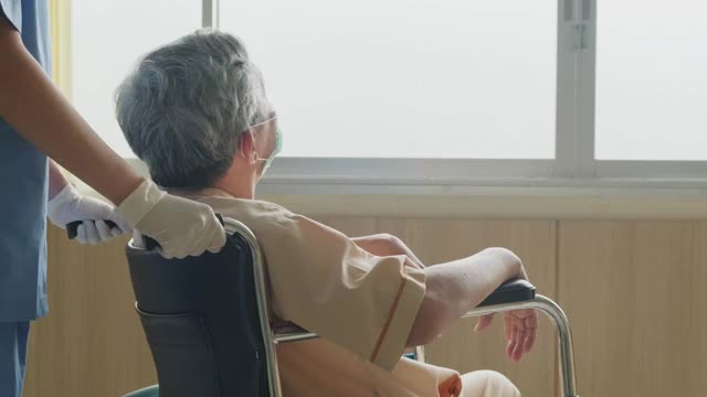 在医院照顾坐在轮椅上的成熟男性病人的亚洲护士。年轻妇女和老人戴上外科口罩，以预防covid - 19大流行。女孩对老人微笑。