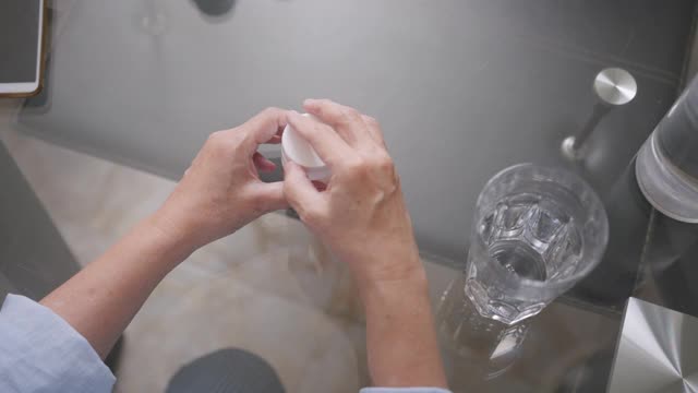 俯视图拍摄的资深亚洲妇女正在吃药或药片。老妇人抱着水喝着药后，坐在厨房的桌子上。
