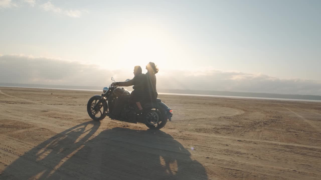 一个男的和一个女的在沙漠里骑摩托车。一对相爱的情侣骑着摩托车在干涸的盐湖上享受日落时分的美好时光。慢动作镜头