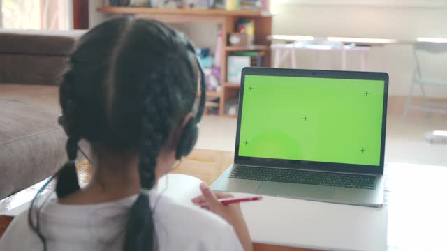亚洲女童学生戴上耳机，通过视频电话与老师在线学习。由于Covid - 19大流行，在隔离期间，孩子正在用电脑笔记本电脑在家学习。