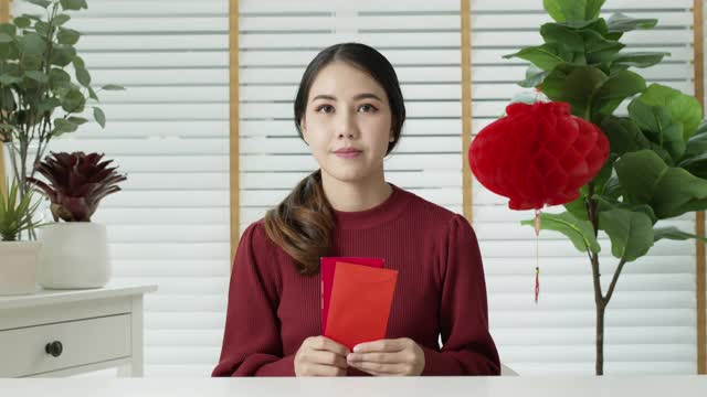 自拍视频通话中，一位迷人的年轻亚洲女士穿着红色的中国新年庆祝服，开心地微笑着交谈，看着拿着红包的手机。