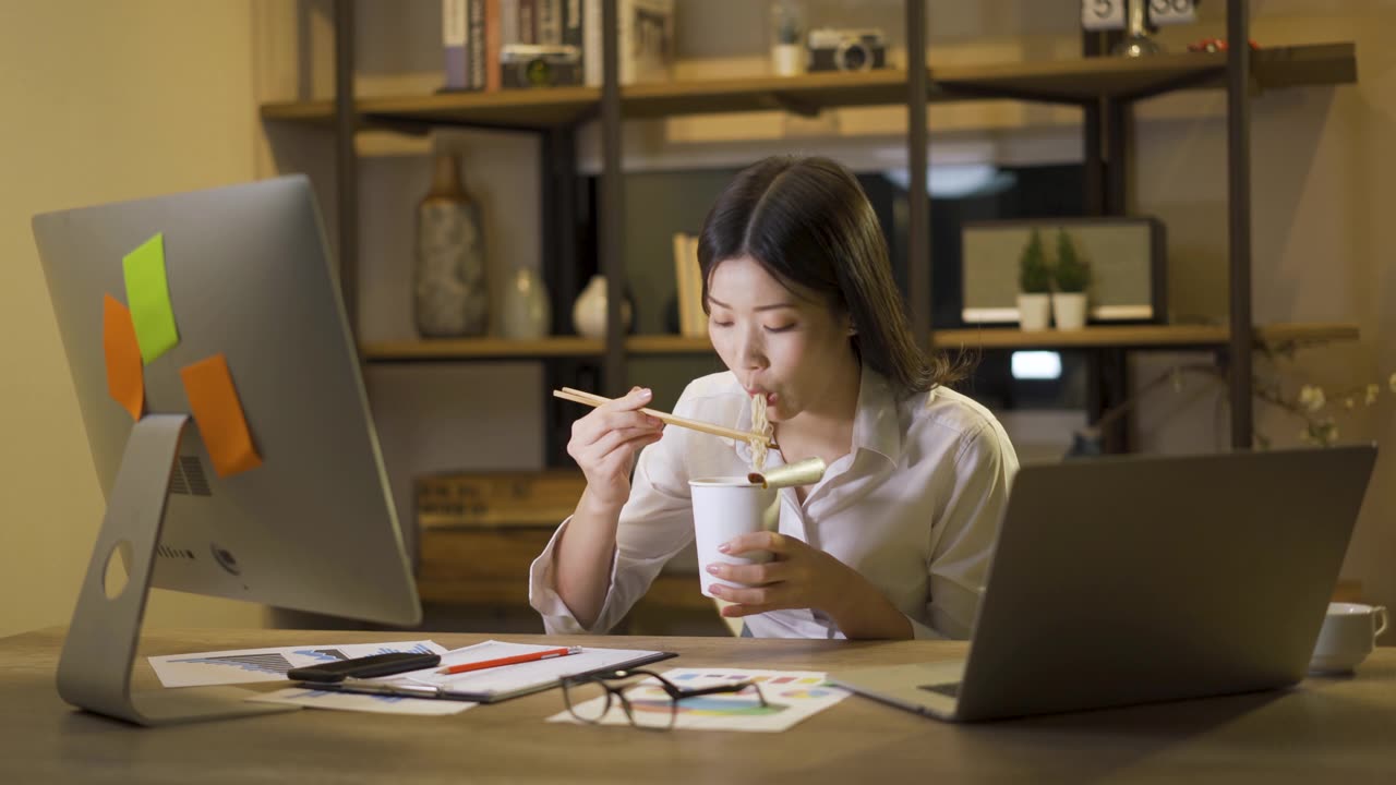 詳情圖-亞洲自由職業(yè)者精明的商業(yè)女性一邊吃著方便面，一邊在家里的客廳里用筆記本電腦工作。