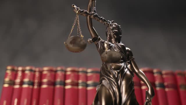 在一所大学的房间里，蒙着眼睛的青铜女神代表着法律雕像旋转着，举着象征公正和两种法律来源的天平和剑，背景是排成一排的红书