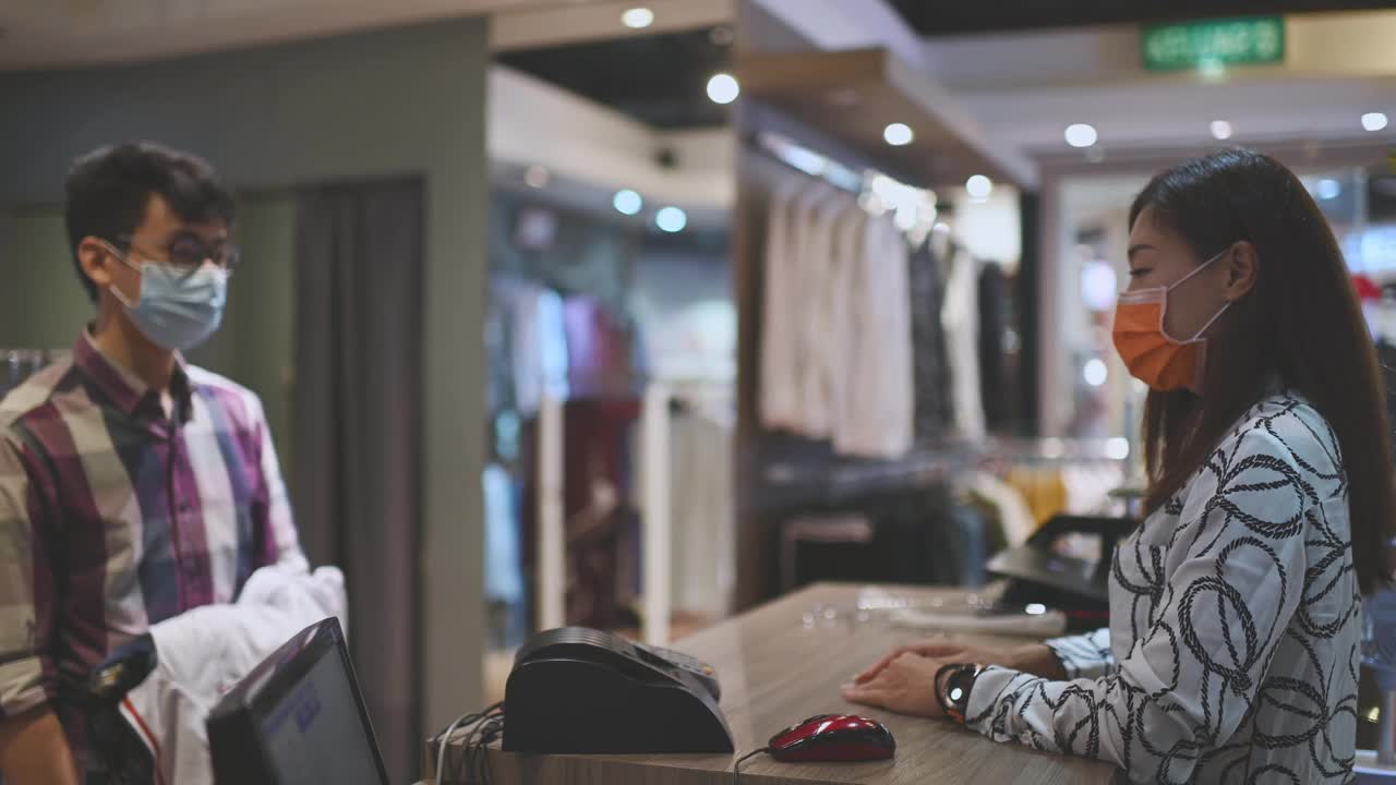 亚洲华人美丽的女男装店店主收银员用口罩折叠衣服，她的顾客把他们放在收银台的袋子里