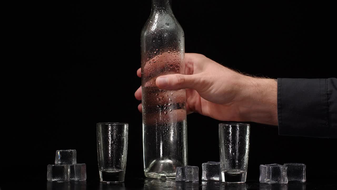 调酒师将伏特加从瓶子中倒入两个带有冰块的小杯中，背景为黑色