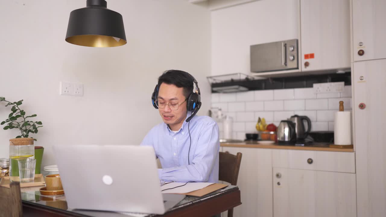 亚洲华人中年男子面带微笑，戴着耳机在用餐区使用笔记本电脑参加在线会议
