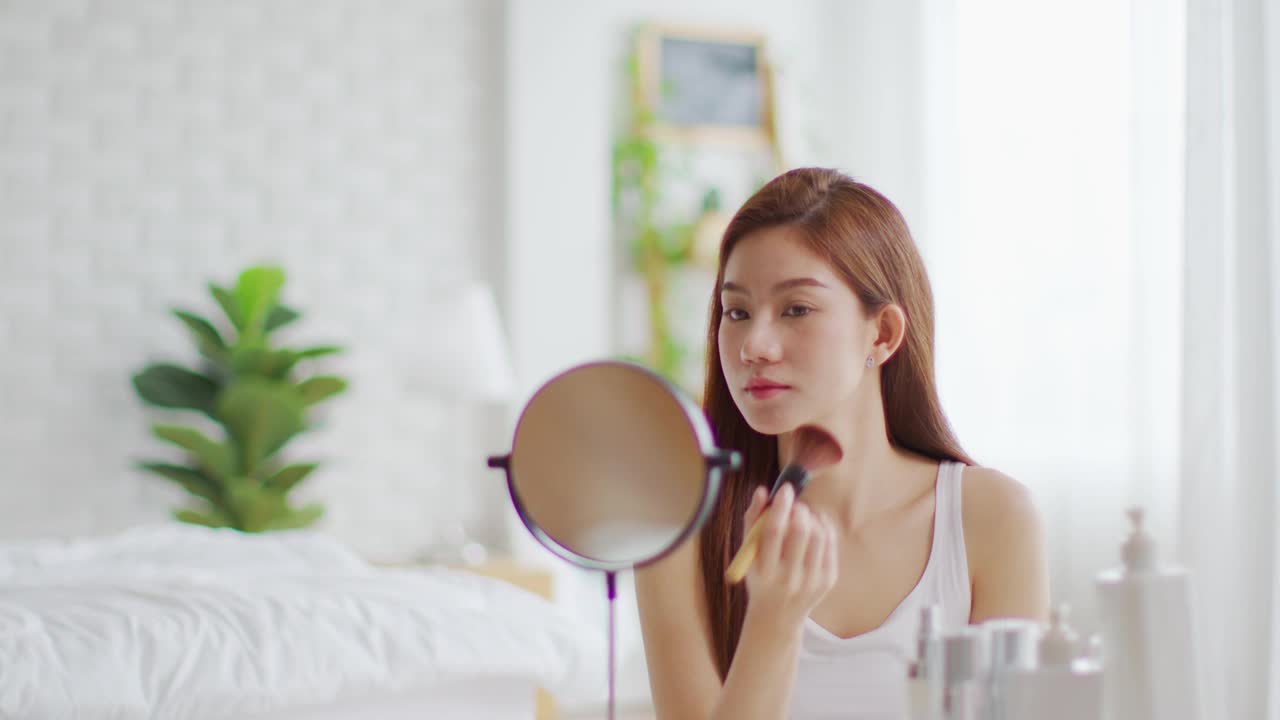 快乐的亚洲年轻女人微笑，在镜子里做皮肤护理，化妆刷在她的脸颊和美容治疗在家，健康女人自我护理与美容产品在家