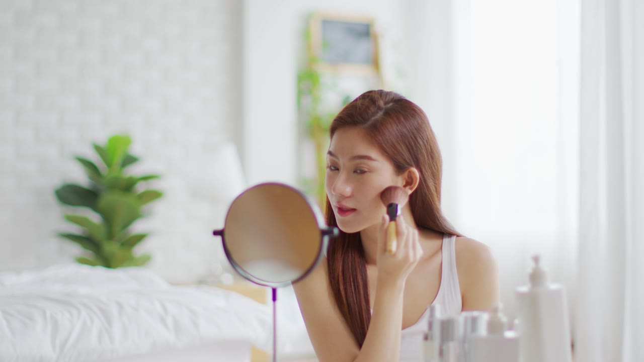 详情图-快乐的亚洲年轻女人微笑，在镜子里做皮肤护理，化妆刷在她的脸颊和美容治疗在家，健康女人自我护理与美容产品在家