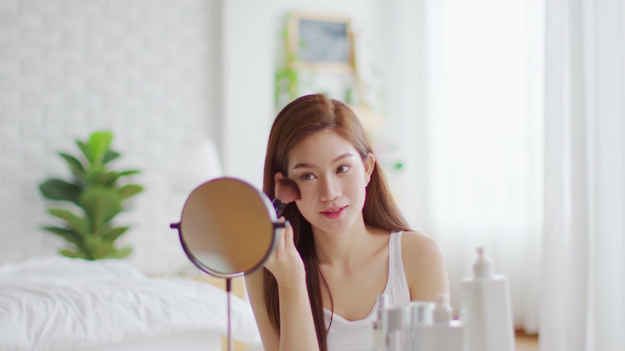 详情图-快乐的亚洲年轻女人微笑，在镜子里做皮肤护理，化妆刷在她的脸颊和美容治疗在家，健康女人自我护理与美容产品在家