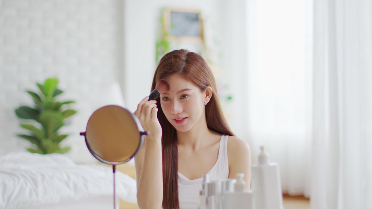 详情图-快乐的亚洲年轻女人微笑，在镜子里做皮肤护理，化妆刷在她的脸颊和美容治疗在家，健康女人自我护理与美容产品在家