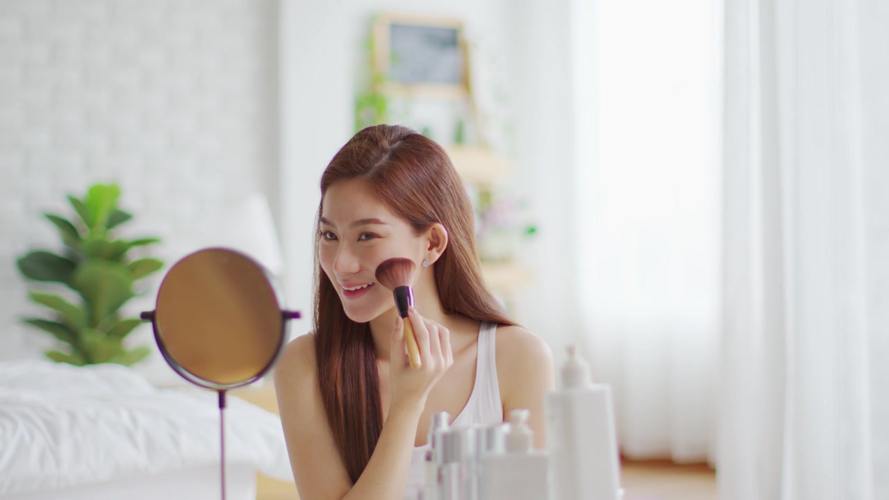 详情图-快乐的亚洲年轻女人微笑，在镜子里做皮肤护理，化妆刷在她的脸颊和美容治疗在家，健康女人自我护理与美容产品在家