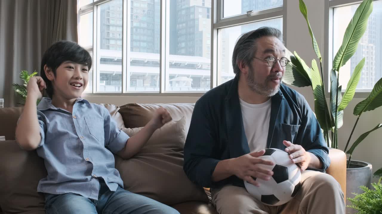 亚洲父亲和儿子在电视上看足球比赛，当球队把球射进球门时，他们高兴地激动起来。