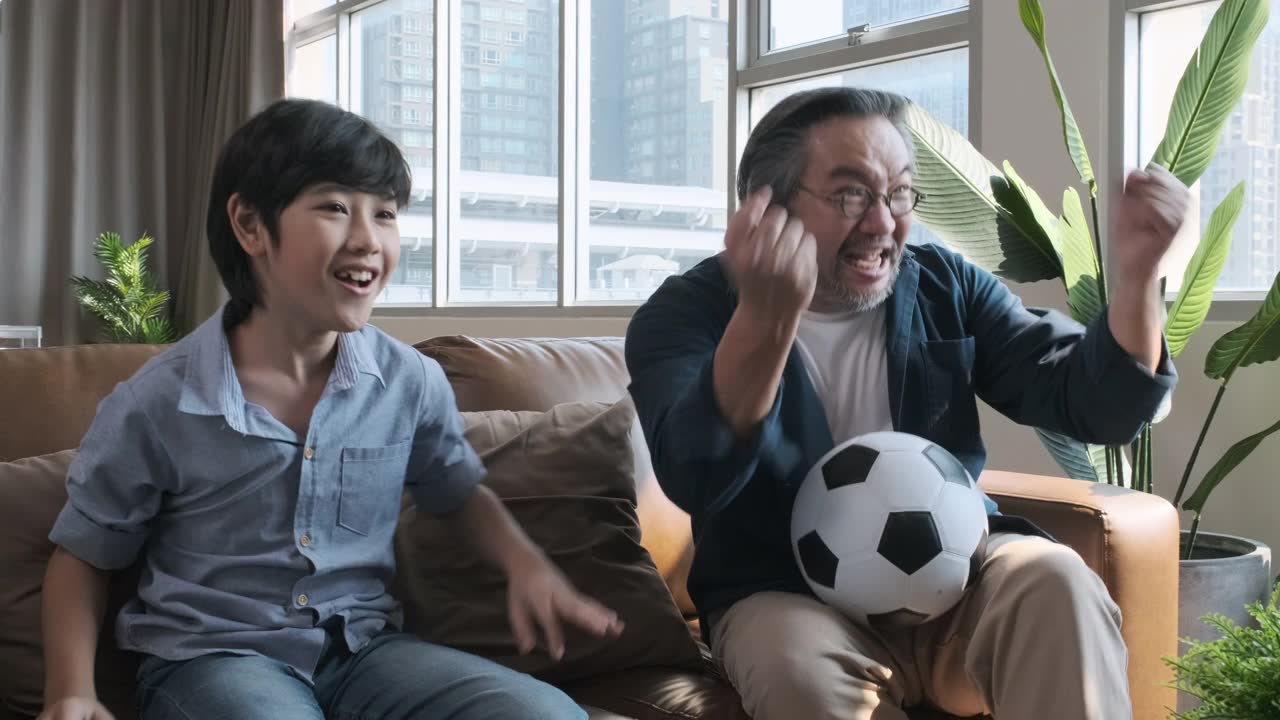 详情图-亚洲父亲和儿子在电视上看足球比赛，当球队把球射进球门时，他们高兴地激动起来。