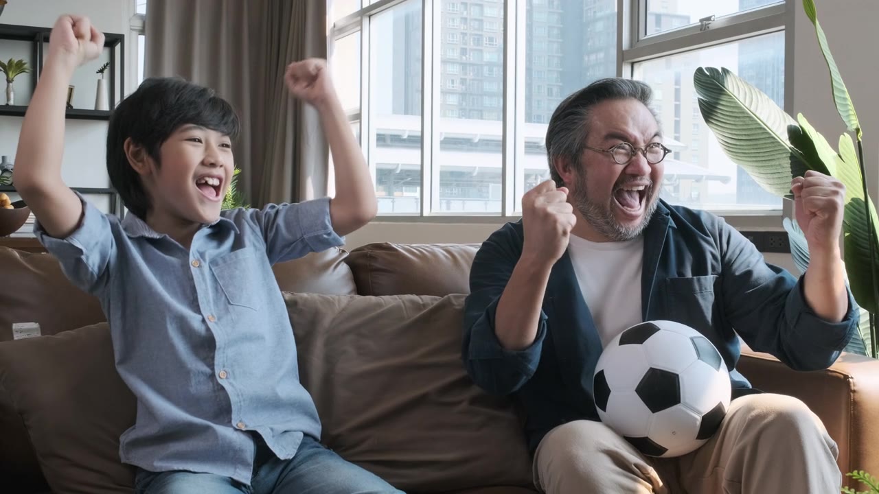 详情图-亚洲父亲和儿子在电视上看足球比赛，当球队把球射进球门时，他们高兴地激动起来。