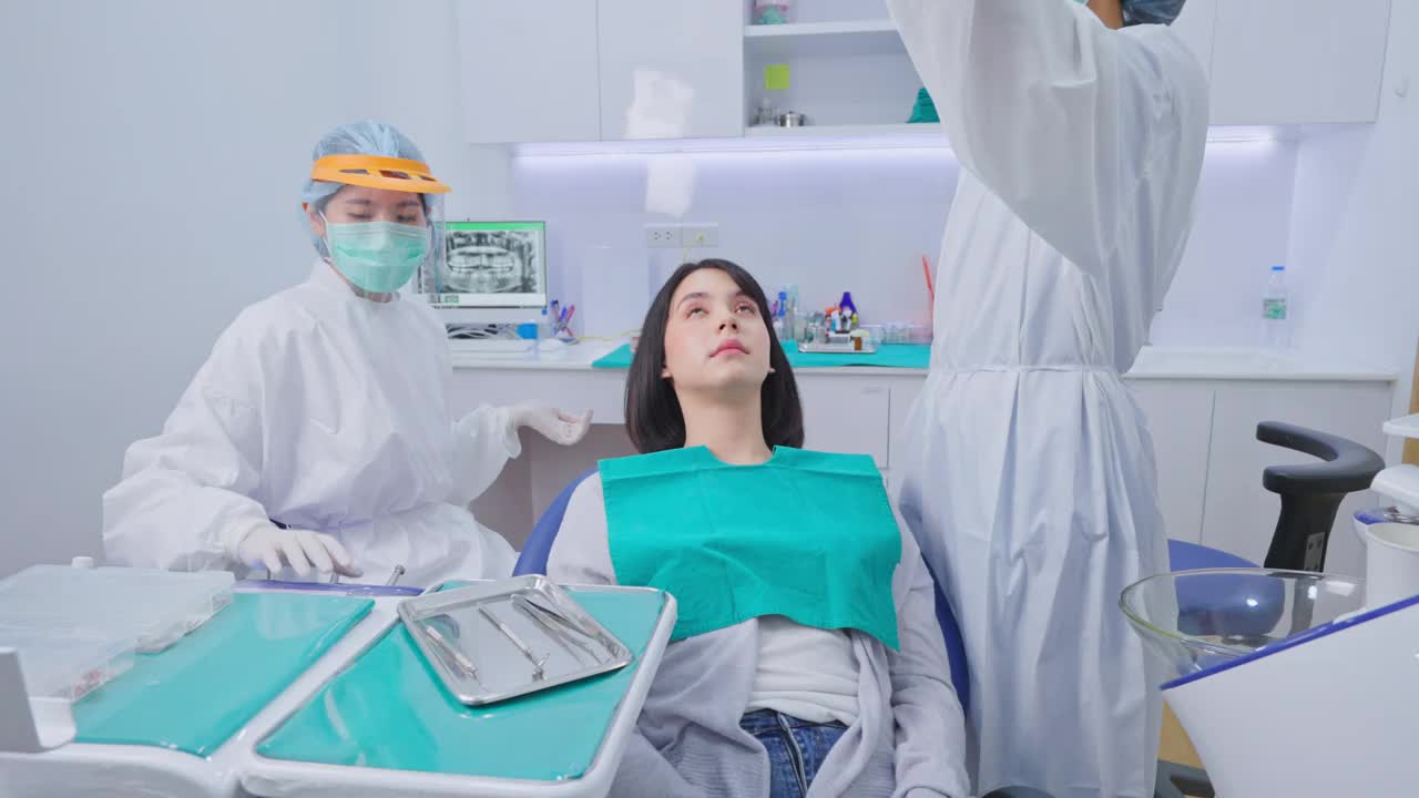 亚洲女牙医穿着实验服，戴着防护面罩和护盾，为小女孩病人调整牙科椅和检查牙齿。协助和调整牙科手术光源