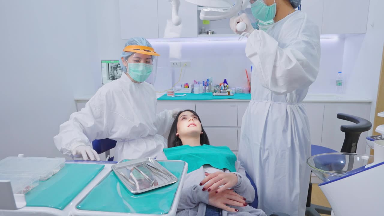 详情图-亚洲女牙医穿着实验服，戴着防护面罩和护盾，为小女孩病人调整牙科椅和检查牙齿。协助和调整牙科手术光源