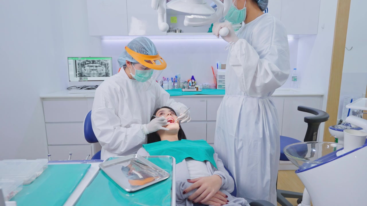 详情图-亚洲女牙医穿着实验服，戴着防护面罩和护盾，为小女孩病人调整牙科椅和检查牙齿。协助和调整牙科手术光源