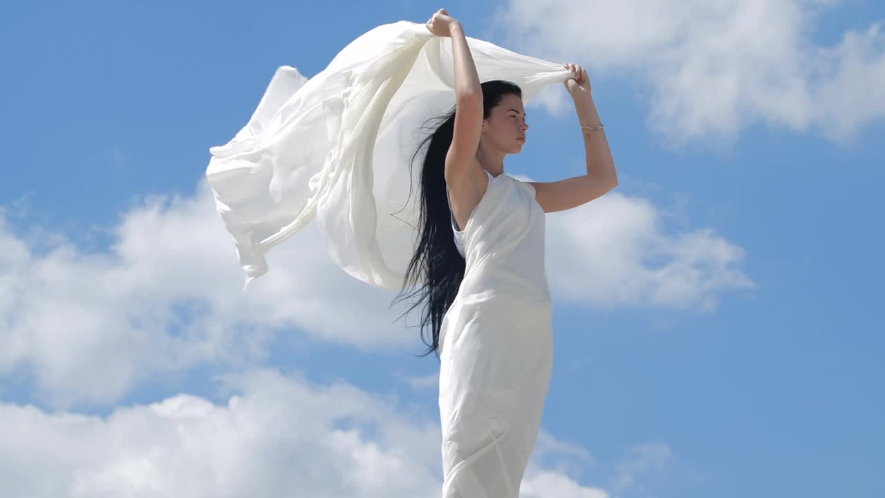 山顶上站着一位美丽的黑发长发女子，她穿着飘逸的白色衣服，映衬着天空。天使存在的幻影。200帧/秒的慢动作