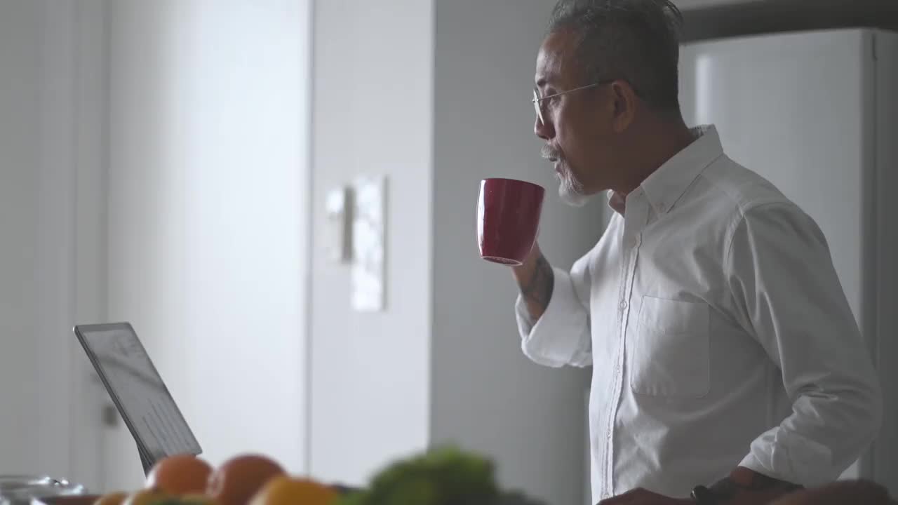 胡子花白头发的亚洲华人老人在厨房柜台边喝着咖啡边用着平板电脑