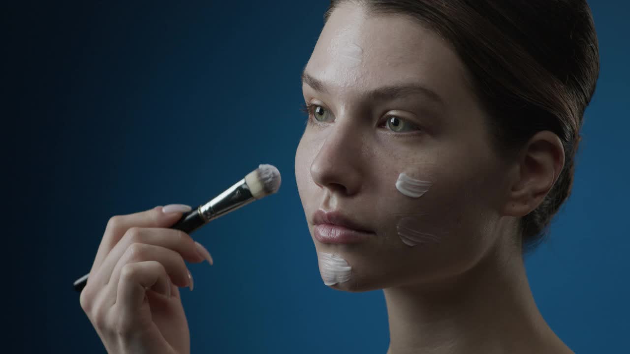 化妆的步骤。一个有着绿色眼睛的黑发女孩将粉底涂抹在脸上，然后用化妆刷将粉底涂抹在皮肤上。