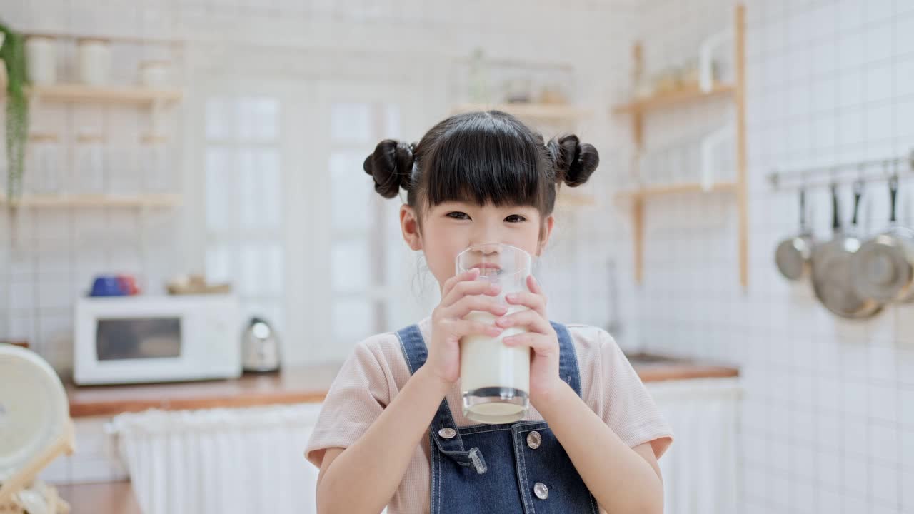 亚洲可爱的小孩拿着一杯牛奶在家里的厨房里喝。学龄前儿童或女儿被牛奶弄脏后，用手臂擦嘴，然后微笑着看镜头。