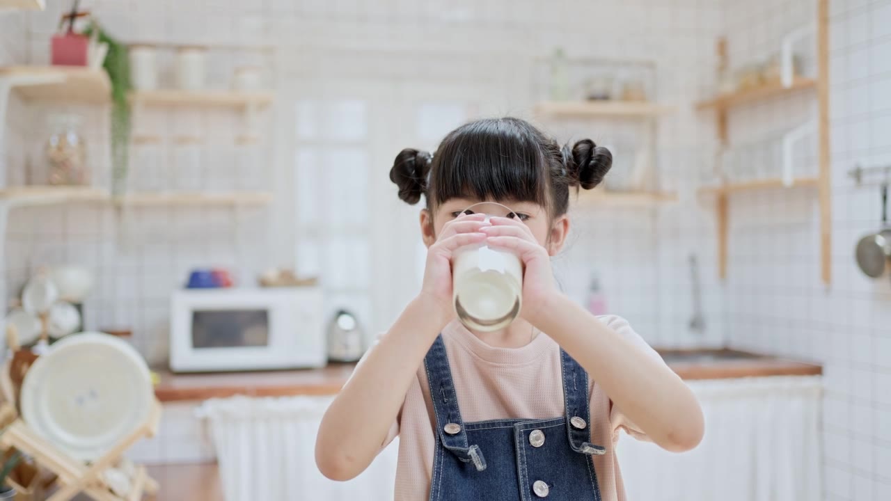 詳情圖-亞洲可愛的小孩拿著一杯牛奶在家里的廚房里喝。學(xué)齡前兒童或女兒被牛奶弄臟后，用手臂擦嘴，然后微笑著看鏡頭。