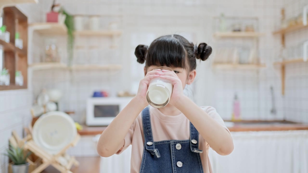 詳情圖-亞洲可愛的小孩拿著一杯牛奶在家里的廚房里喝。學(xué)齡前兒童或女兒被牛奶弄臟后，用手臂擦嘴，然后微笑著看鏡頭。