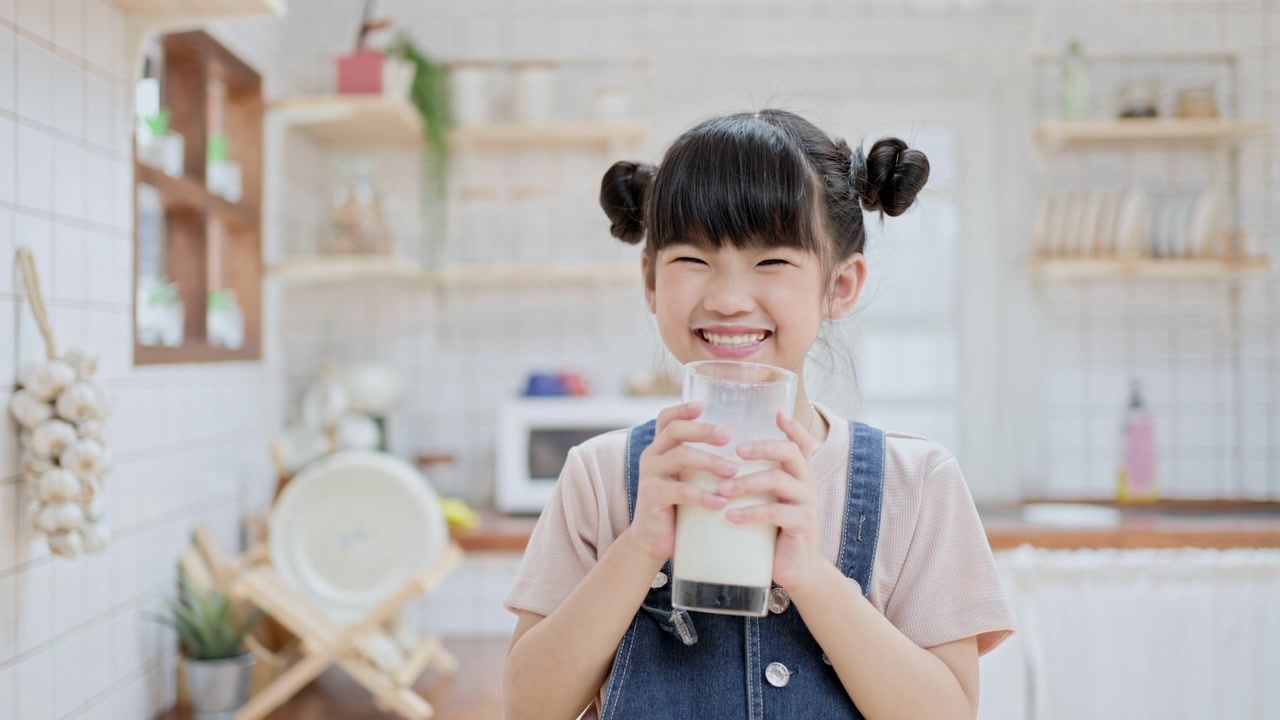 詳情圖-亞洲可愛的小孩拿著一杯牛奶在家里的廚房里喝。學(xué)齡前兒童或女兒被牛奶弄臟后，用手臂擦嘴，然后微笑著看鏡頭。